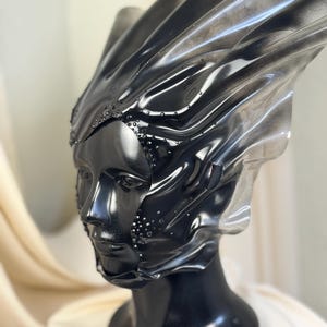 Könnte beinhalten: Ein schwarzer und silberner Kopfschmuck mit einem geformten Gesicht und einem fließenden, geschichteten Design. Der Kopfschmuck ist mit kleinen, funkelnden Verzierungen versehen und auf einem schwarzen Schaufensterpuppenkopf montiert. Die Gesamtästhetik ist futuristisch und avantgardistisch.
