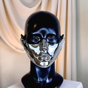 Op de afbeelding: Een zwarte mannequinhoofd met een glanzend zilveren gezichtsmasker. Het masker bedekt de neus, mond en wangen, met ingewikkelde geometrische ontwerpen. De mannequin staat op een wit oppervlak, met een crèmekleurig gordijn op de achtergrond.