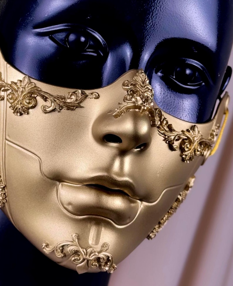 K&ouml;nnte beinhalten: Eine goldfarbene Maske f&uuml;r Maskenb&auml;lle mit aufwendigen, erhabenen floralen und scrollartigen Details. Die Maske bedeckt das untere Gesicht und weist eine modellierte Nase und Lippen auf. Die Maske wird auf einem schwarzen Schaufensterpuppenkopf pr&auml;sentiert.