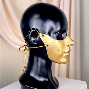 K&ouml;nnte beinhalten: Eine goldfarbene Maske mit Halbgesichtsdesign, mit Nase und Mund, wird auf einem schwarzen Schaufensterkopf pr&auml;sentiert. Die Maske ist mit einem goldenen Faden befestigt. Die B&uuml;ste steht auf einer wei&szlig;en Oberfl&auml;che.