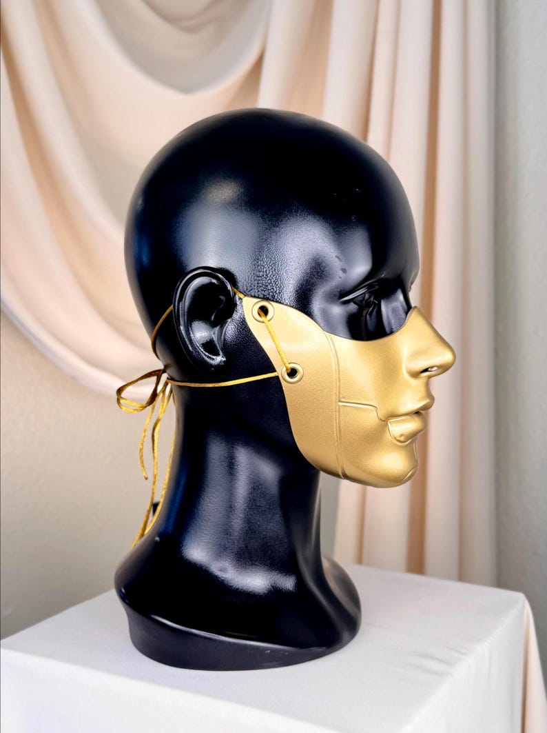 K&ouml;nnte beinhalten: Eine goldfarbene Maske mit Halbgesichtsdesign, mit Nase und Mund, wird auf einem schwarzen Schaufensterkopf pr&auml;sentiert. Die Maske ist mit einem goldenen Faden befestigt. Die B&uuml;ste steht auf einer wei&szlig;en Oberfl&auml;che.