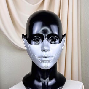 May include: A futuristic silver mask with a detailed design, displayed on a black mannequin head. The mask covers the nose and mouth, with intricate lines and shapes. The mannequin is on a white surface, with a cream-colored curtain backdrop.