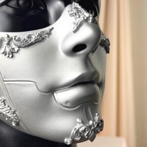 K&ouml;nnte beinhalten: Silberne Maske f&uuml;r Maskenb&auml;lle mit erhabenen floralen Details. Die Maske bedeckt Nase und Mund, vor schwarzem Hintergrund. Die Maske hat eine glatte, matte Oberfl&auml;che. Entworfen f&uuml;r eine formelle Veranstaltung oder ein Kost&uuml;m.