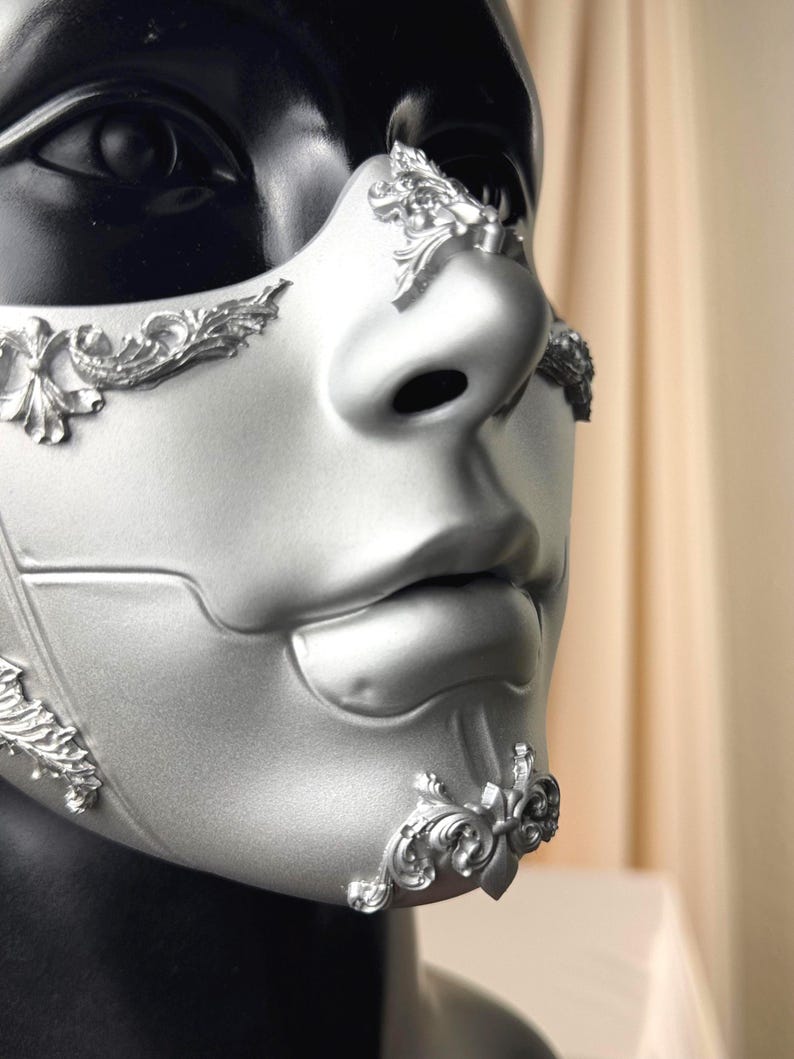 K&ouml;nnte beinhalten: Silberne Maske f&uuml;r Maskenb&auml;lle mit erhabenen floralen Details. Die Maske bedeckt Nase und Mund, vor schwarzem Hintergrund. Die Maske hat eine glatte, matte Oberfl&auml;che. Entworfen f&uuml;r eine formelle Veranstaltung oder ein Kost&uuml;m.