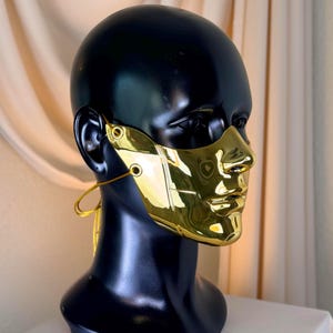 K&ouml;nnte beinhalten: Eine goldfarbene Gesichtsmaske mit reflektierendem, metallischem Finish. Die Maske ist so konzipiert, dass sie Nase und Mund bedeckt und ein skulpturales Aussehen hat. Sie wird auf einem schwarzen Schaufensterpuppenkopf mit einer goldfarbenen Schnur am Ohr pr&auml;sentiert.