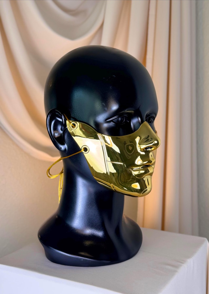 K&ouml;nnte beinhalten: Eine goldfarbene Gesichtsmaske mit reflektierendem, metallischem Finish. Die Maske ist so konzipiert, dass sie Nase und Mund bedeckt und ein skulpturales Aussehen hat. Sie wird auf einem schwarzen Schaufensterpuppenkopf mit einer goldfarbenen Schnur am Ohr pr&auml;sentiert.