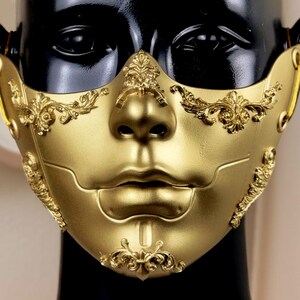 K&ouml;nnte beinhalten: Eine goldfarbene Maske mit aufwendigen Verzierungen im Barockstil. Die Maske bedeckt das untere Gesicht und weist eine modellierte Nase und Lippen auf. Dekorative Verzierungen schm&uuml;cken die Wangen und die Stirn und schaffen eine luxuri&ouml;se und theatralische &Auml;sthetik. Die Maske wird auf einem schwarzen Schaufensterkopf pr&auml;sentiert.