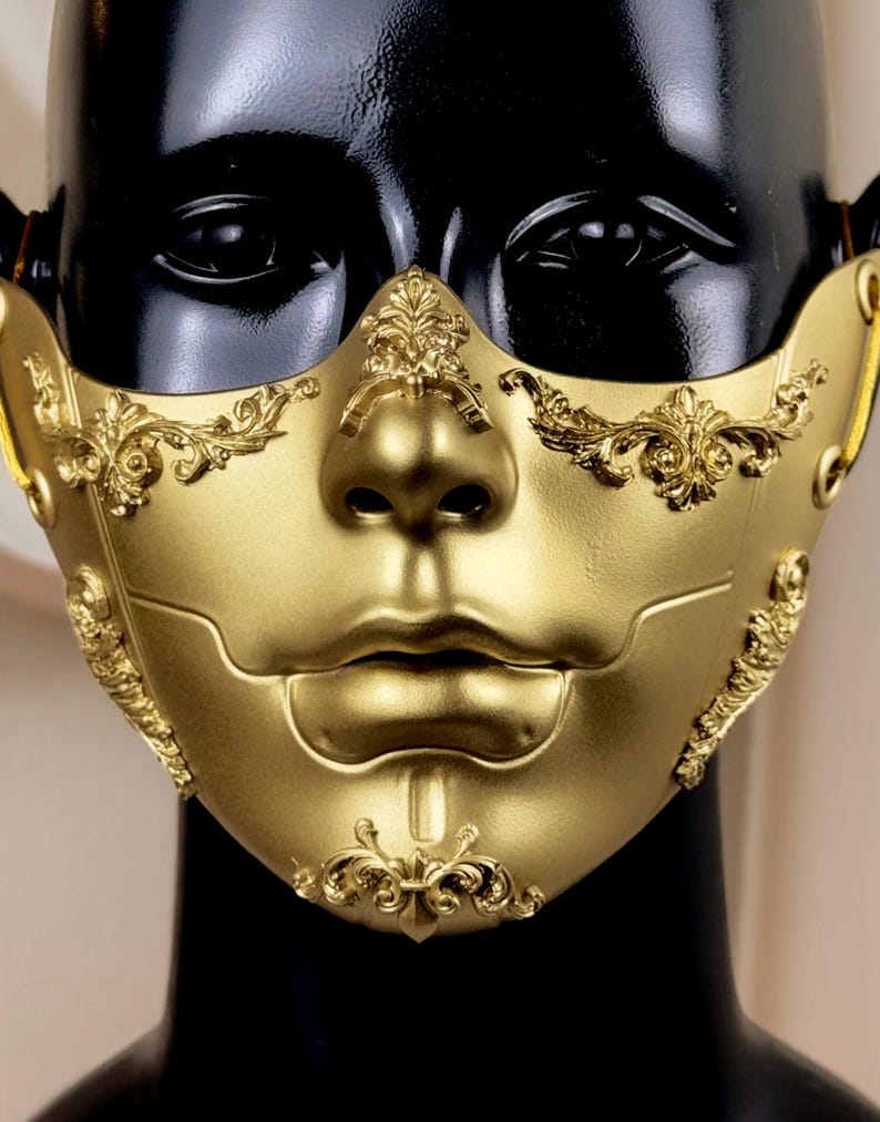 K&ouml;nnte beinhalten: Eine goldfarbene Maske mit aufwendigen Verzierungen im Barockstil. Die Maske bedeckt das untere Gesicht und weist eine modellierte Nase und Lippen auf. Dekorative Verzierungen schm&uuml;cken die Wangen und die Stirn und schaffen eine luxuri&ouml;se und theatralische &Auml;sthetik. Die Maske wird auf einem schwarzen Schaufensterkopf pr&auml;sentiert.