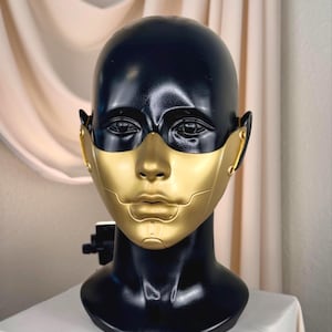 K&ouml;nnte beinhalten: Eine schwarz-goldene Maske auf einem schwarzen Schaufensterkopf. Die Maske hat ein goldenes Gesicht und einen schwarzen Kopf. Die Maske ist mit einer futuristischen &Auml;sthetik gestaltet. Der Schaufensterkopf steht auf einer wei&szlig;en Oberfl&auml;che.
