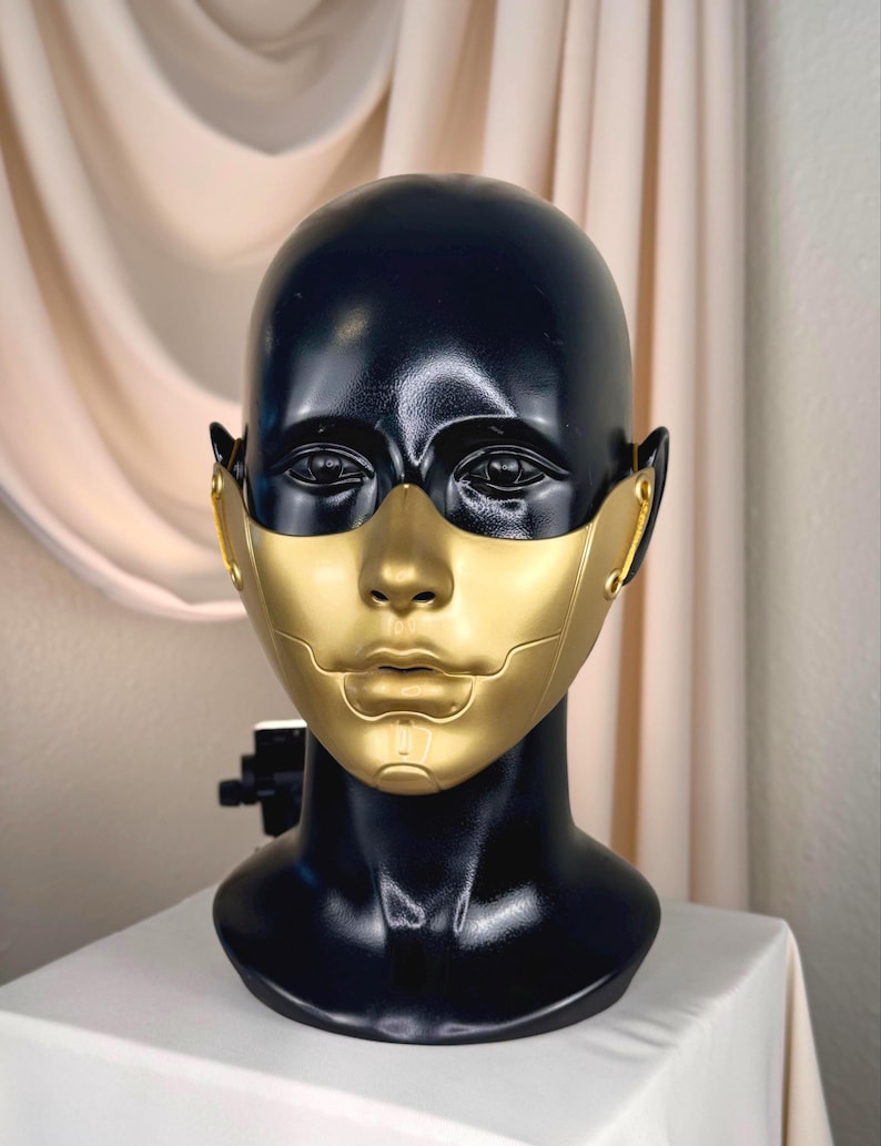K&ouml;nnte beinhalten: Eine schwarz-goldene Maske auf einem schwarzen Schaufensterkopf. Die Maske hat ein goldenes Gesicht und einen schwarzen Kopf. Die Maske ist mit einer futuristischen &Auml;sthetik gestaltet. Der Schaufensterkopf steht auf einer wei&szlig;en Oberfl&auml;che.