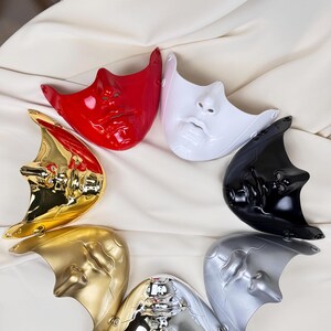 K&ouml;nnte beinhalten: Eine Sammlung von sieben Gesichtsmasken in verschiedenen Farben, darunter Rot, Wei&szlig;, Schwarz, Silber, Gold und Chrom. Die Masken sind kreisf&ouml;rmig auf einem wei&szlig;en Stoffhintergrund angeordnet. Jede Maske zeigt ein stilisiertes menschliches Gesicht.
