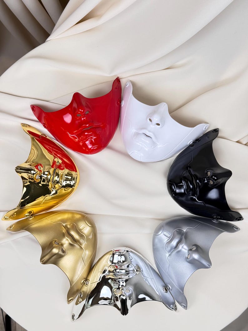 K&ouml;nnte beinhalten: Eine Sammlung von sieben Gesichtsmasken in verschiedenen Farben, darunter Rot, Wei&szlig;, Schwarz, Silber, Gold und Chrom. Die Masken sind kreisf&ouml;rmig auf einem wei&szlig;en Stoffhintergrund angeordnet. Jede Maske zeigt ein stilisiertes menschliches Gesicht.