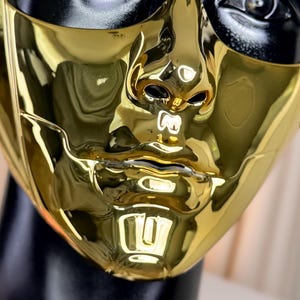 K&ouml;nnte beinhalten: Nahaufnahme einer goldfarbenen Maske mit reflektierender Oberfl&auml;che. Die Maske hat ein menschliches Gesichtsdesign mit Ausschnitten f&uuml;r Nase, Mund und Augen. Die Oberseite der Maske ist schwarz. Die Maske befindet sich auf einem schwarzen Schaufensterkopf.