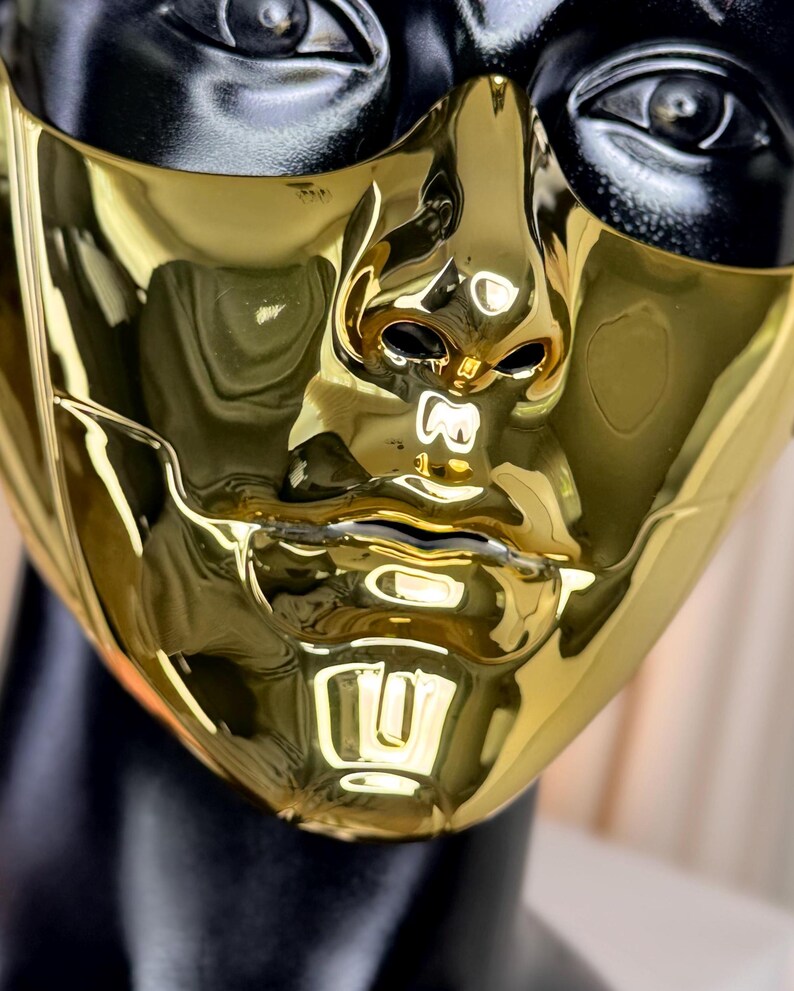 K&ouml;nnte beinhalten: Nahaufnahme einer goldfarbenen Maske mit reflektierender Oberfl&auml;che. Die Maske hat ein menschliches Gesichtsdesign mit Ausschnitten f&uuml;r Nase, Mund und Augen. Die Oberseite der Maske ist schwarz. Die Maske befindet sich auf einem schwarzen Schaufensterkopf.