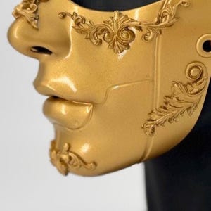K&ouml;nnte beinhalten: Eine goldfarbene Maske mit aufwendigen, erhabenen floralen Designs. Die Maske bedeckt Nase und Mund und hat ein skulpturales Aussehen. Die Maske hat eine gl&auml;nzende Oberfl&auml;che und besteht wahrscheinlich aus einem haltbaren Material, das f&uuml;r Kost&uuml;me oder Dekorationszwecke geeignet ist.