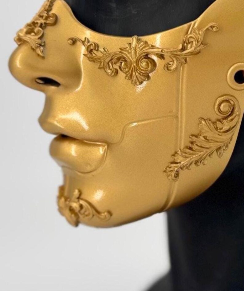 K&ouml;nnte beinhalten: Eine goldfarbene Maske mit aufwendigen, erhabenen floralen Designs. Die Maske bedeckt Nase und Mund und hat ein skulpturales Aussehen. Die Maske hat eine gl&auml;nzende Oberfl&auml;che und besteht wahrscheinlich aus einem haltbaren Material, das f&uuml;r Kost&uuml;me oder Dekorationszwecke geeignet ist.