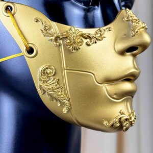 K&ouml;nnte beinhalten: Eine goldfarbene Maske mit aufwendigen, erhabenen floralen Designs. Die Maske bedeckt Nase und Mund und hat &Ouml;sen zur Befestigung. Sie wird auf einem schwarzen Schaufensterkopf pr&auml;sentiert, um ihre komplizierten Details und Handwerkskunst zu zeigen.