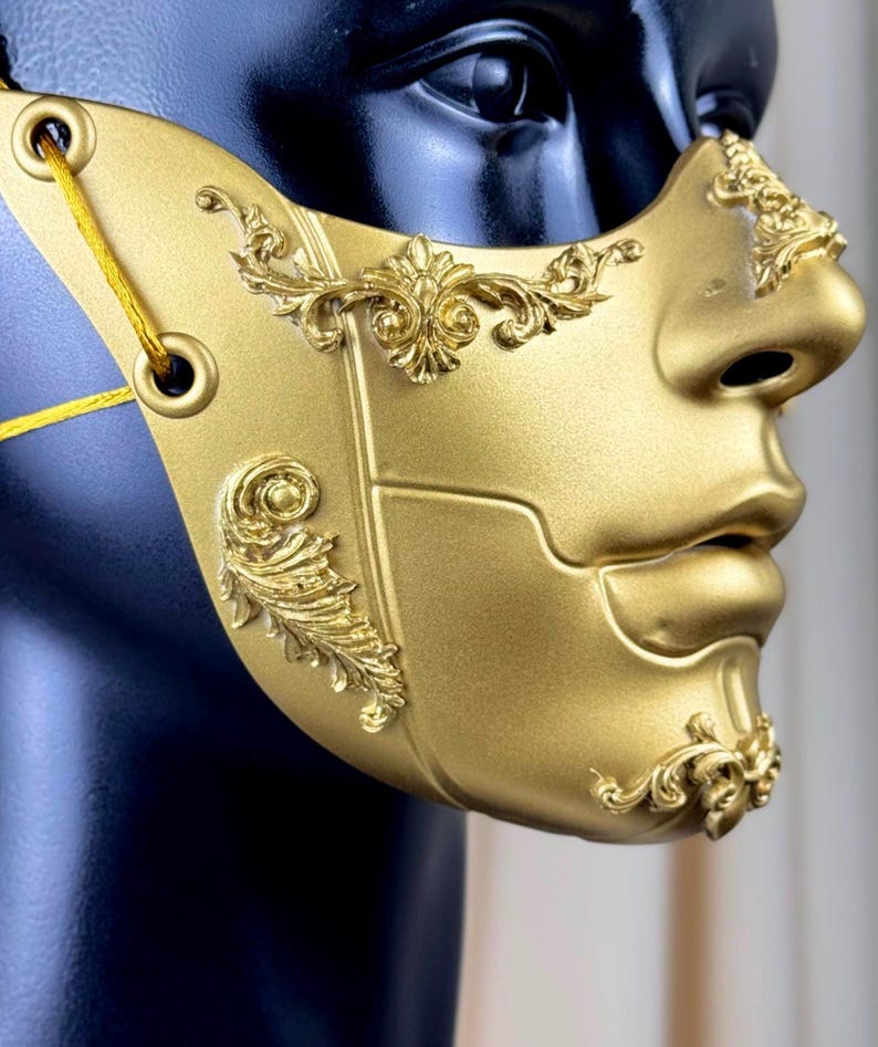 K&ouml;nnte beinhalten: Eine goldfarbene Maske mit aufwendigen, erhabenen floralen Designs. Die Maske bedeckt Nase und Mund und hat &Ouml;sen zur Befestigung. Sie wird auf einem schwarzen Schaufensterkopf pr&auml;sentiert, um ihre komplizierten Details und Handwerkskunst zu zeigen.