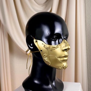 K&ouml;nnte beinhalten: Eine goldfarbene Halbmaske mit aufwendigen, klassisch anmutenden Details. Die Maske wird auf einem schwarzen Schaufensterkopf pr&auml;sentiert, um ihr Design und ihre Form hervorzuheben. Die Maske hat einen metallischen Glanz und wird mit d&uuml;nnen goldenen Kordeln befestigt.