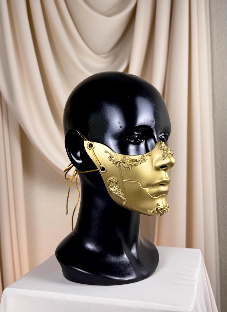 K&ouml;nnte beinhalten: Eine goldfarbene Halbmaske mit aufwendigen, klassisch anmutenden Details. Die Maske wird auf einem schwarzen Schaufensterkopf pr&auml;sentiert, um ihr Design und ihre Form hervorzuheben. Die Maske hat einen metallischen Glanz und wird mit d&uuml;nnen goldenen Kordeln befestigt.