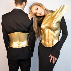 K&ouml;nnte beinhalten: Zwei Personen in schwarzen Outfits mit goldfarbener K&ouml;rperpanzerung. Eine Person tr&auml;gt einen Anzug mit goldenem G&uuml;rtel, die andere ein Kleid mit goldener Brustplatte. Die goldenen Teile weisen detaillierte Designs auf.