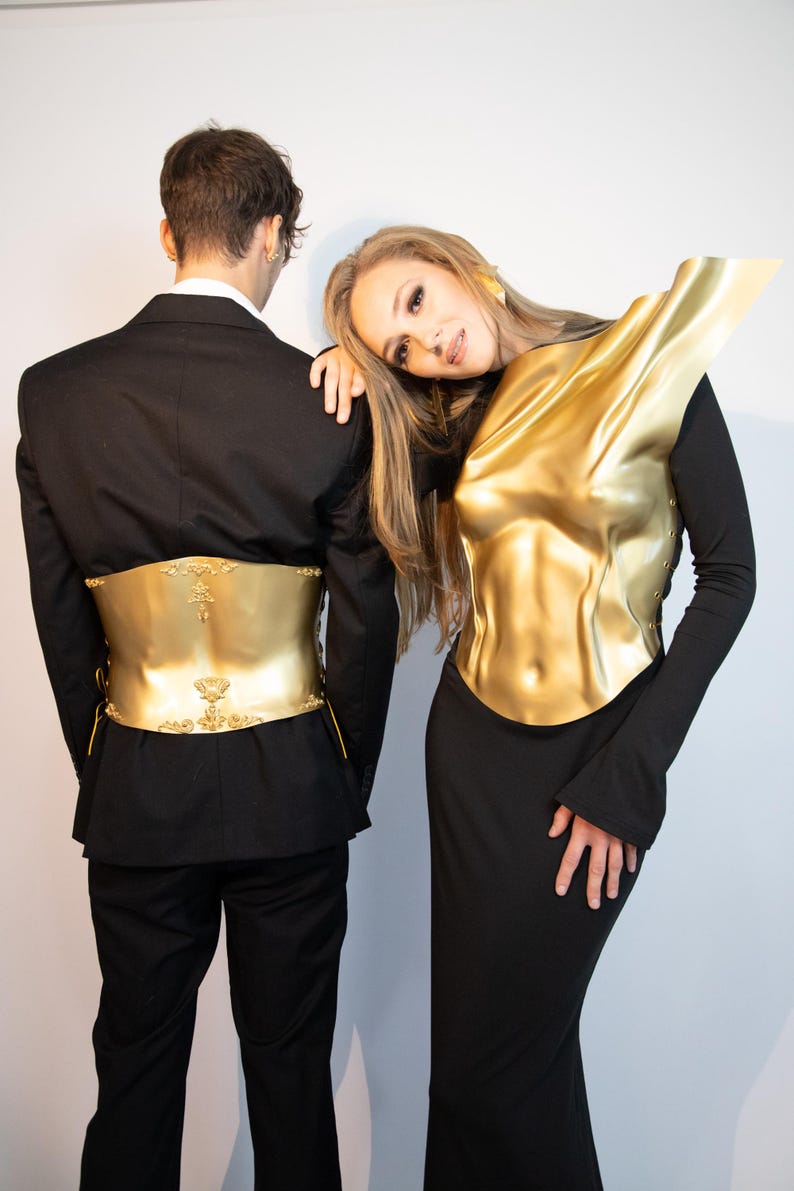 K&ouml;nnte beinhalten: Zwei Personen in schwarzen Outfits mit goldfarbener K&ouml;rperpanzerung. Eine Person tr&auml;gt einen Anzug mit goldenem G&uuml;rtel, die andere ein Kleid mit goldener Brustplatte. Die goldenen Teile weisen detaillierte Designs auf.