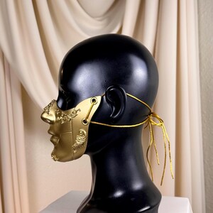 K&ouml;nnte beinhalten: Eine goldfarbene Maske mit aufwendigen Details, pr&auml;sentiert auf einem schwarzen Schaufensterkopf. Die Maske bedeckt Nase und Untergesicht und wird mit goldenem Faden befestigt. Der Hintergrund zeigt einen cremefarbenen, drapierten Stoff.