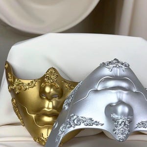 K&ouml;nnte beinhalten: Zwei dekorative Masken, eine goldene und eine silberne, liegen auf einer wei&szlig;en Oberfl&auml;che. Die Masken weisen Gesichtsdetails und aufwendige Verzierungen im klassischen Stil auf. Die goldene Maske hat einen warmen, metallischen Glanz, w&auml;hrend die silberne Maske ein k&uuml;hles, mattes Finish hat.