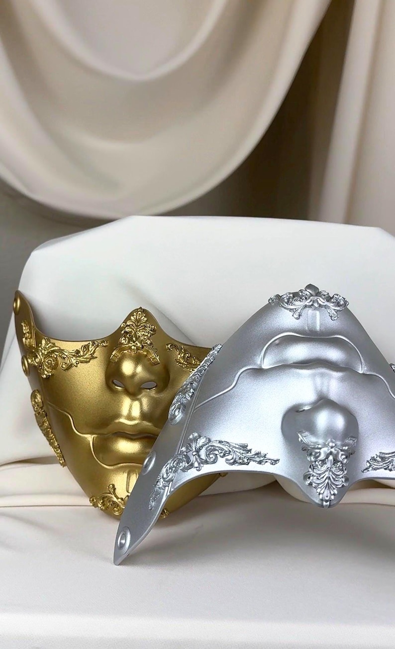 K&ouml;nnte beinhalten: Zwei dekorative Masken, eine goldene und eine silberne, liegen auf einer wei&szlig;en Oberfl&auml;che. Die Masken weisen Gesichtsdetails und aufwendige Verzierungen im klassischen Stil auf. Die goldene Maske hat einen warmen, metallischen Glanz, w&auml;hrend die silberne Maske ein k&uuml;hles, mattes Finish hat.