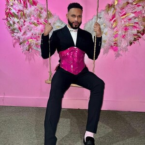 K&ouml;nnte beinhalten: Eine Person tr&auml;gt einen schwarzen Blazer, eine schwarze Hose und ein leuchtend pinkfarbenes Korsett. Die Person sitzt auf einer Holzschaukel mit dekorativen Engelsfl&uuml;geln im Hintergrund. Der Hintergrund ist einfarbig rosa.