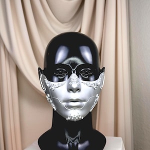 K&ouml;nnte beinhalten: Eine silberne und schwarze Maske f&uuml;r Maskenb&auml;lle mit aufwendigen Details. Die Maske wird auf einem schwarzen Schaufensterkopf pr&auml;sentiert, um ihr Design hervorzuheben. Der silberne Teil bedeckt das untere Gesicht mit dekorativen Akzenten.