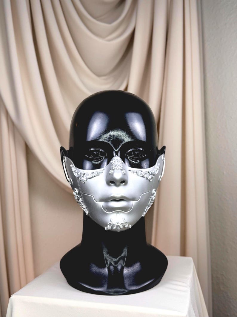 K&ouml;nnte beinhalten: Eine silberne und schwarze Maske f&uuml;r Maskenb&auml;lle mit aufwendigen Details. Die Maske wird auf einem schwarzen Schaufensterkopf pr&auml;sentiert, um ihr Design hervorzuheben. Der silberne Teil bedeckt das untere Gesicht mit dekorativen Akzenten.