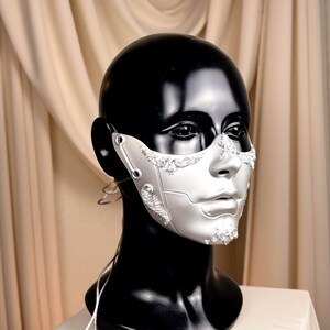 K&ouml;nnte beinhalten: Silberne und wei&szlig;e Maske f&uuml;r Maskenb&auml;lle mit aufwendigen Details. Die Maske wird auf einem schwarzen Schaufensterpuppenkopf pr&auml;sentiert, um ihr Design hervorzuheben. Die Maske hat komplizierte Muster und ein metallisches Finish, ideal f&uuml;r Kost&uuml;mpartys oder Theaterveranstaltungen.