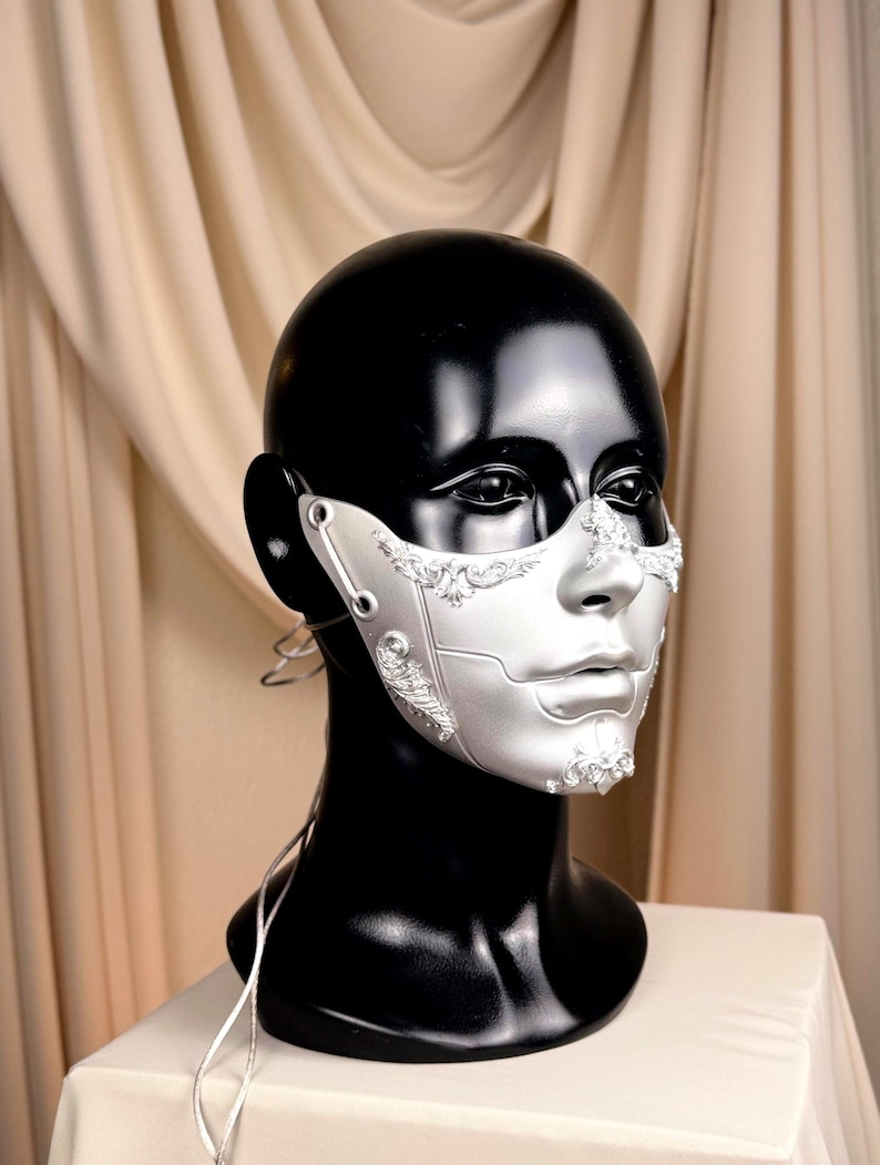 K&ouml;nnte beinhalten: Silberne und wei&szlig;e Maske f&uuml;r Maskenb&auml;lle mit aufwendigen Details. Die Maske wird auf einem schwarzen Schaufensterpuppenkopf pr&auml;sentiert, um ihr Design hervorzuheben. Die Maske hat komplizierte Muster und ein metallisches Finish, ideal f&uuml;r Kost&uuml;mpartys oder Theaterveranstaltungen.