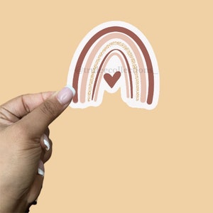 May include: A sticker of a rainbow with a heart in the center. The rainbow is made of pink, brown, and beige with gold glitter accents.