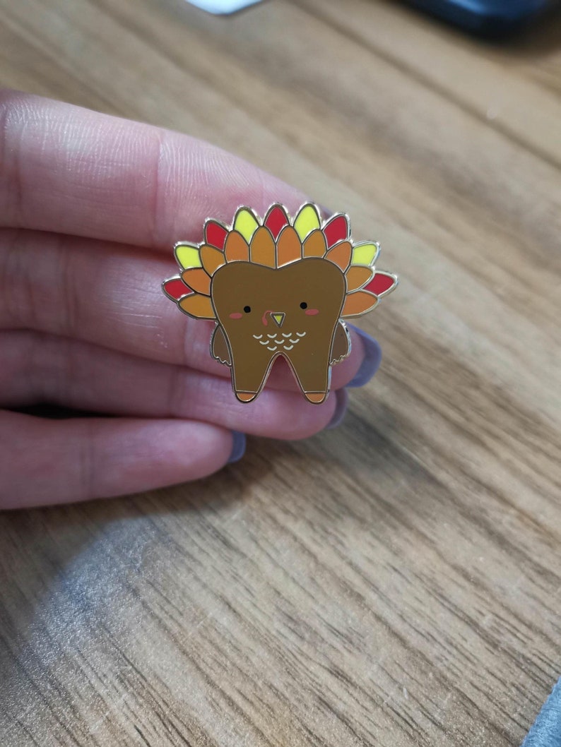 May include: A gold-colored enamel pin shaped like a tooth with a turkey design. The tooth has a brown body, a red beak, and a yellow and orange feathered head.