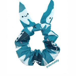May include: Teal blue scrunchie with a white tooth pattern. The teeth have a smiling face. The scrunchie is tied in a bow with the ends of the bow shaped like teeth. The text "@dentaldiy" is printed on the bottom right corner of the scrunchie.