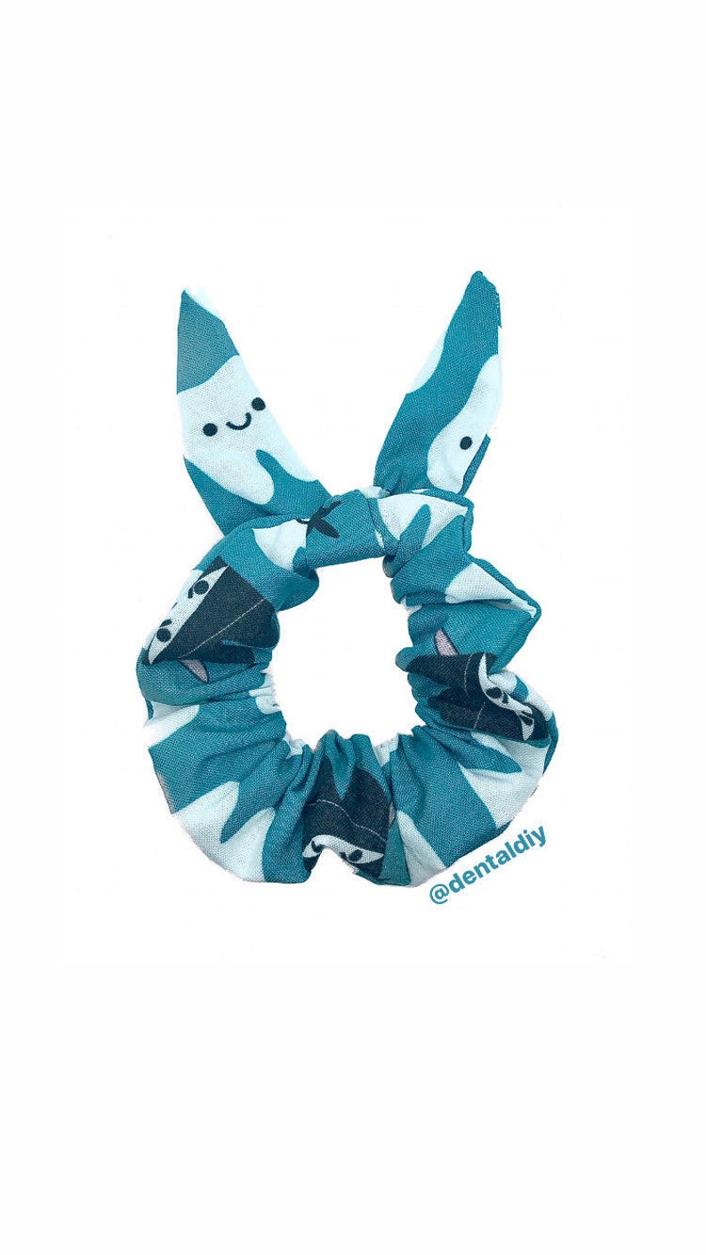May include: Teal blue scrunchie with a white tooth pattern. The teeth have a smiling face. The scrunchie is tied in a bow with the ends of the bow shaped like teeth. The text "@dentaldiy" is printed on the bottom right corner of the scrunchie.