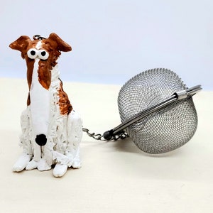 May include: A whimsical tea infuser featuring a seated dog figurine with brown and white coloring. The dog is attached by a chain to a stainless steel mesh tea ball, perfect for steeping loose leaf tea.