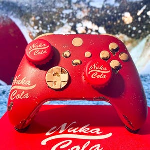 Può includere: Controller di gioco rosso con il logo Nuka Cola e dettagli di tappi di bottiglia. Il controller è su una superficie rossa con il logo Nuka Cola. Il design ha un'estetica retrò e vintage.