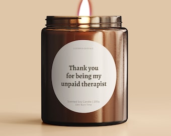 May include: A brown glass candle with a white label that reads "Thank you for being my unpaid therapist". The candle is lit and has a flame.