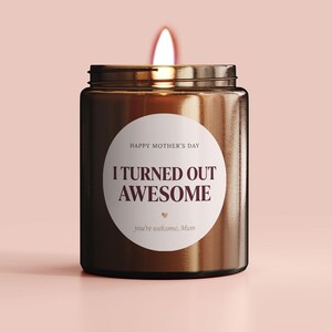 May include: A lit brown glass candle with a metal lid. The candle has a white circular label with the text "HAPPY MOTHER'S DAY" above "I TURNED OUT AWESOME" and "you're welcome, Mum" below. The candle is on a pink background.