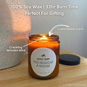 May include: A brown glass candle jar with a white label that reads "HOLY SHIT YOU BOUGHT A HOUSE". The candle has a crackling wooden wick and a 33-hour burn time. The candle is on a wooden surface with a black lid next to it.