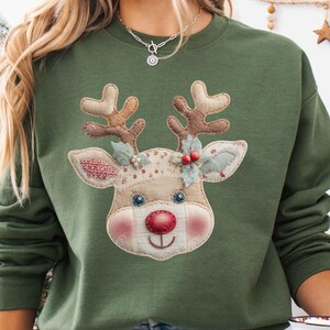 Peut inclure: Un sweat-shirt vert olive avec un motif de visage de renne, un nez rouge et des bois bruns. Le sweat-shirt est porté avec un jean bleu clair. Une tasse blanche avec un motif de Père Noël est tenue à la main.