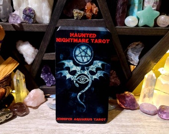 Pode incluir: Uma carta de tarô com o título "Haunted Nightmare Tarot" em texto vermelho. A carta apresenta um fundo azul escuro com um pentagrama branco e um design de olho estilizado. O texto "Jennifer Aquarius Tarot" está na parte inferior.
