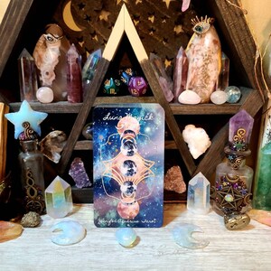 K&ouml;nnte beinhalten: Ein h&ouml;lzernes Regal mit dreieckigem Design, gef&uuml;llt mit Kristallen, Tarotkarten und dekorativen Flaschen. Die Tarotkarte zeigt ein himmlisches Design mit dem Text "Luna Magick" und "Jennifer Aquarius Tarot."