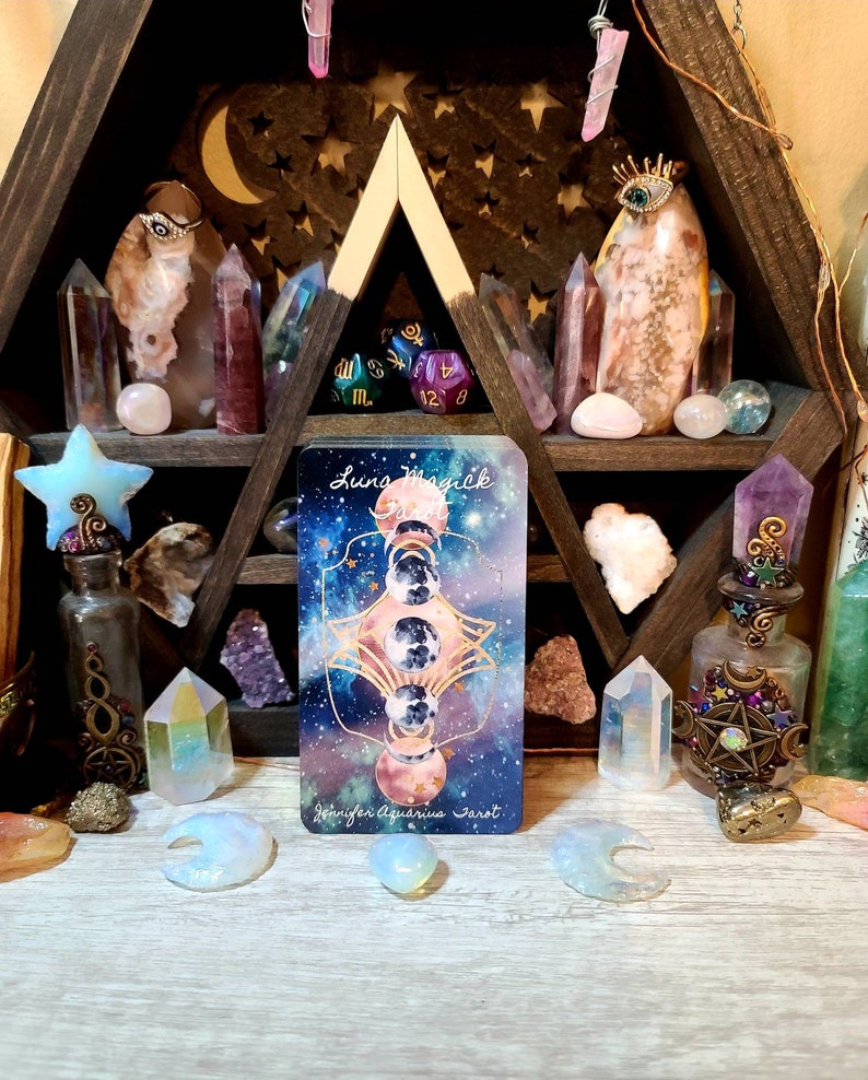 K&ouml;nnte beinhalten: Ein h&ouml;lzernes Regal mit dreieckigem Design, gef&uuml;llt mit Kristallen, Tarotkarten und dekorativen Flaschen. Die Tarotkarte zeigt ein himmlisches Design mit dem Text "Luna Magick" und "Jennifer Aquarius Tarot."