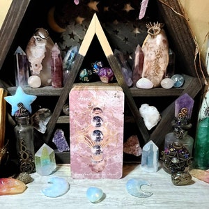K&ouml;nnte beinhalten: Ein sechseckiges Holzregal mit Kristallen, Tarotkarten und dekorativen Flaschen. Die Tarotkarte in der Mitte tr&auml;gt den Text "Luna Moon Tarot" und zeigt Mondphasen. Verschiedene Kristalle in Rosa, Lila und Klar sind angeordnet, was eine mystische Atmosph&auml;re schafft.