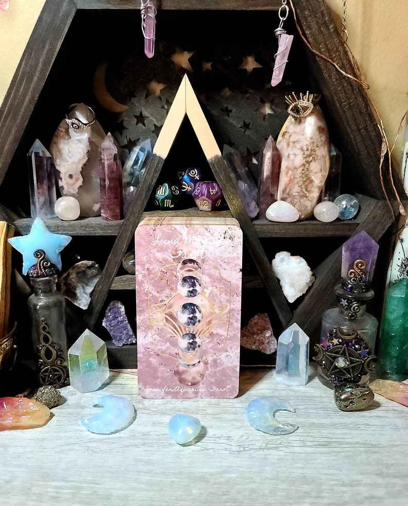 K&ouml;nnte beinhalten: Ein sechseckiges Holzregal mit Kristallen, Tarotkarten und dekorativen Flaschen. Die Tarotkarte in der Mitte tr&auml;gt den Text "Luna Moon Tarot" und zeigt Mondphasen. Verschiedene Kristalle in Rosa, Lila und Klar sind angeordnet, was eine mystische Atmosph&auml;re schafft.