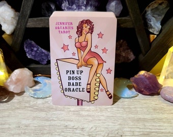 May include: A pink and yellow oracle card featuring a cartoon illustration of a woman in a pink dress sitting on a sign that reads "Pin Up Boss Babe Oracle". The card has a pink background with white stars and the text "Jennifer Aquarius Tarot" at the top.